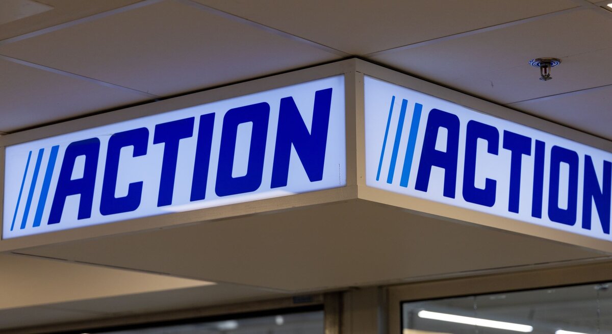 Leuchtendes Aktions-Schild mit blauem Schriftzug „ACTION“ und drei Streifen hängt unter weißer Decke im Eingangsbereich.