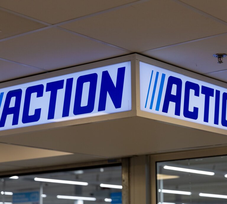 Leuchtendes Aktions-Schild mit blauem Schriftzug „ACTION“ und drei Streifen hängt unter weißer Decke im Eingangsbereich. | © AdobeStock/PixelBiss