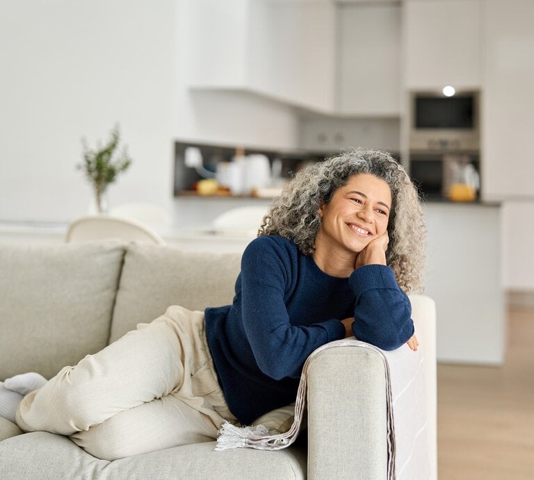 Eine grauhaarige Frau mittleren Alters liegt entspannt lächelnd in heller Jogginghose und dunkelblauem Pullover auf einem Sofa. | © stock.adobe.com/insta_photos