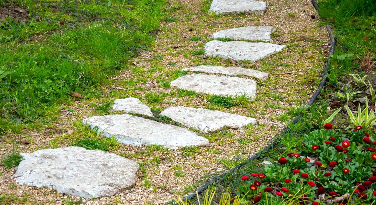 Unregelmäßig geformte Trittsteine führen geschwungen über einen Kiesweg durch einen gepflegten Garten mit frischem Grün.