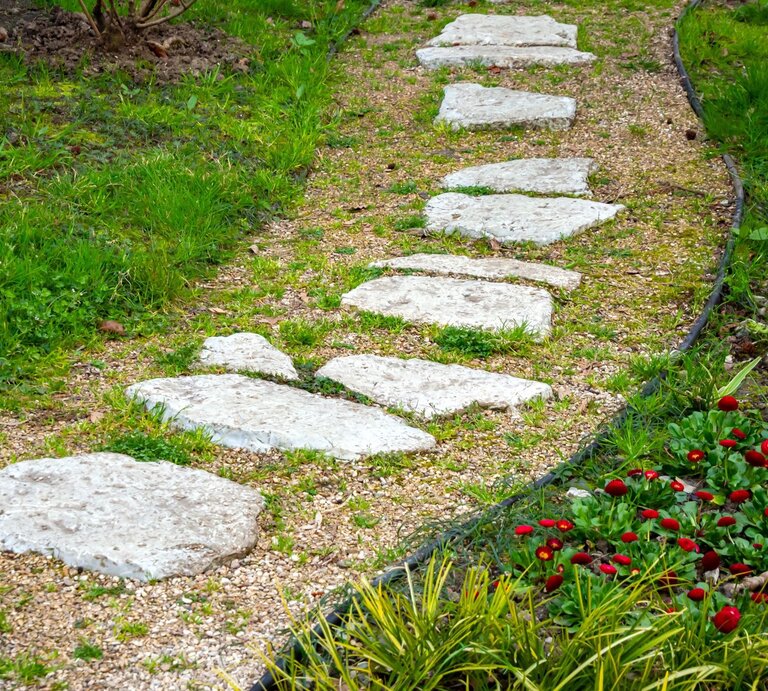 Unregelmäßig geformte Trittsteine führen geschwungen über einen Kiesweg durch einen gepflegten Garten mit frischem Grün. | © Shutterstock/VPales