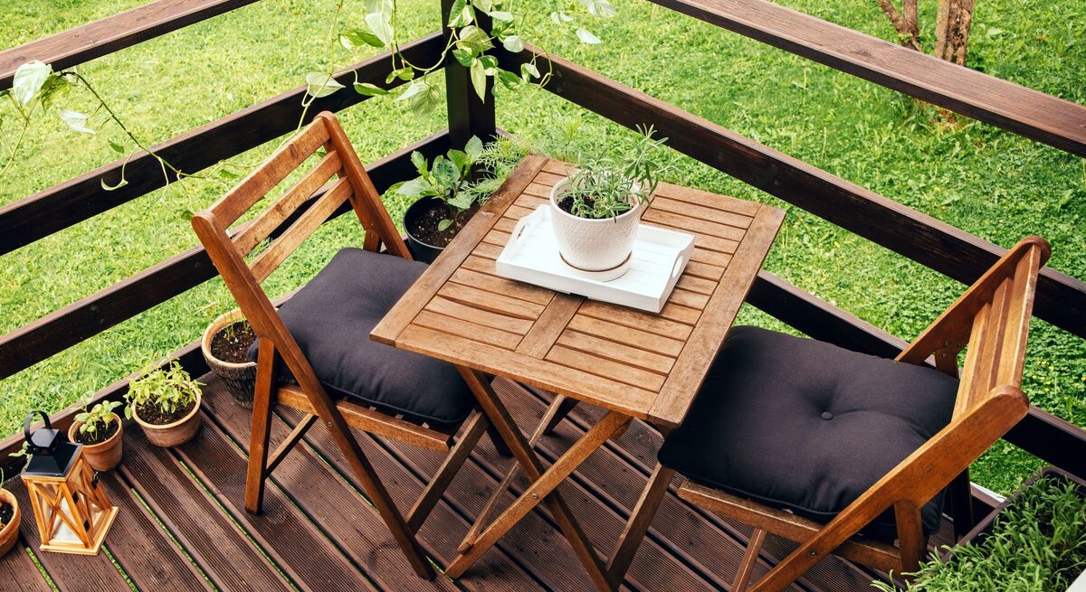 Zwei Holzklappstühle mit schwarzen Polstern stehen an einem kleinen Holztisch auf einer Terrasse mit Kräutertöpfen und Grünpflanzen.