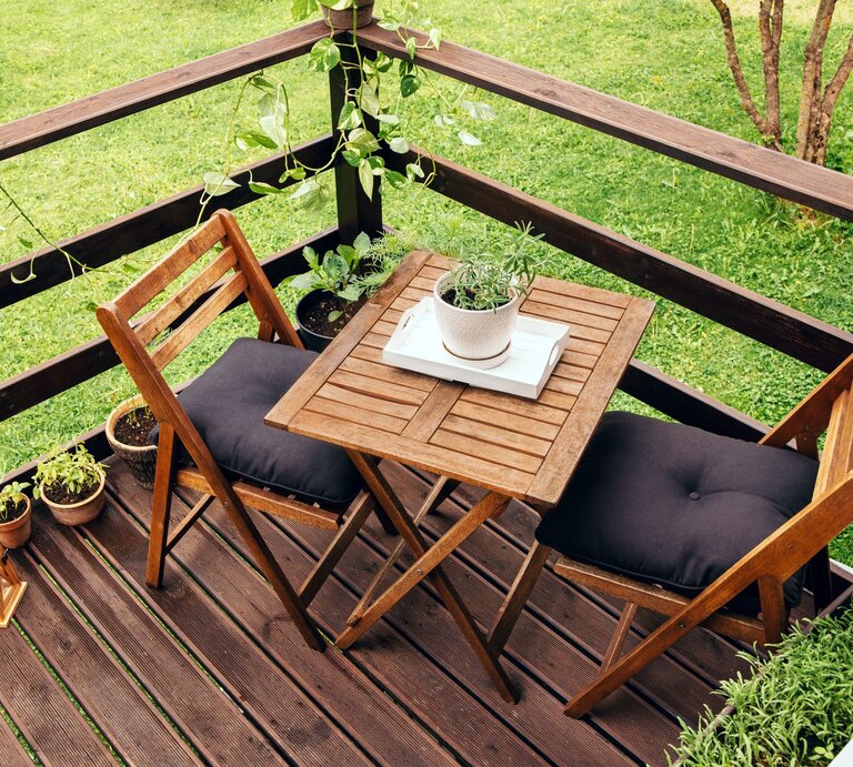 Zwei Holzklappstühle mit schwarzen Polstern stehen an einem kleinen Holztisch auf einer Terrasse mit Kräutertöpfen und Grünpflanzen. | © Shutterstock/FotoHelin