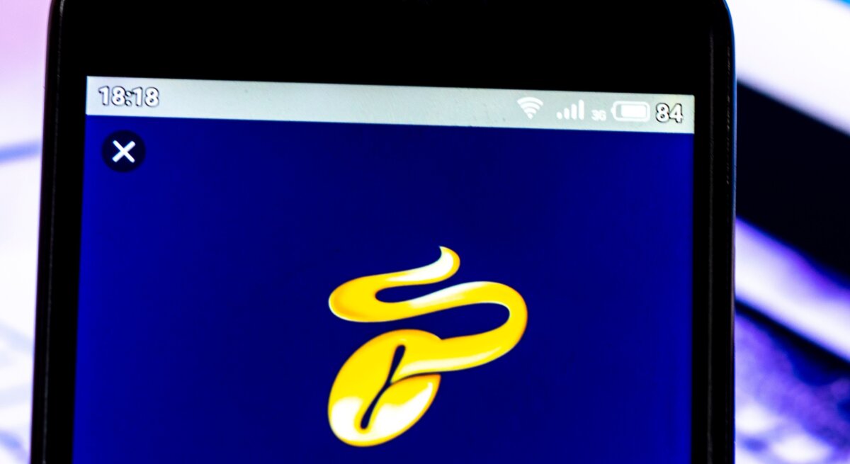 Tchibo-Logo auf einem Smartphone-Bildschirm vor einem unscharfen Laptop, der einen Kursverlauf anzeigt.