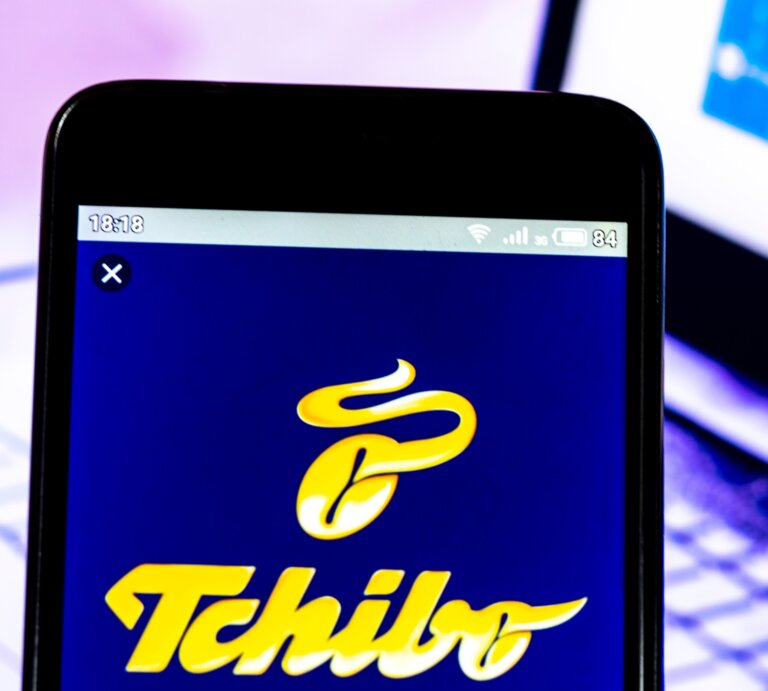 Tchibo-Logo auf einem Smartphone-Bildschirm vor einem unscharfen Laptop, der einen Kursverlauf anzeigt. | © AdobeStock/Игорь Головнёв