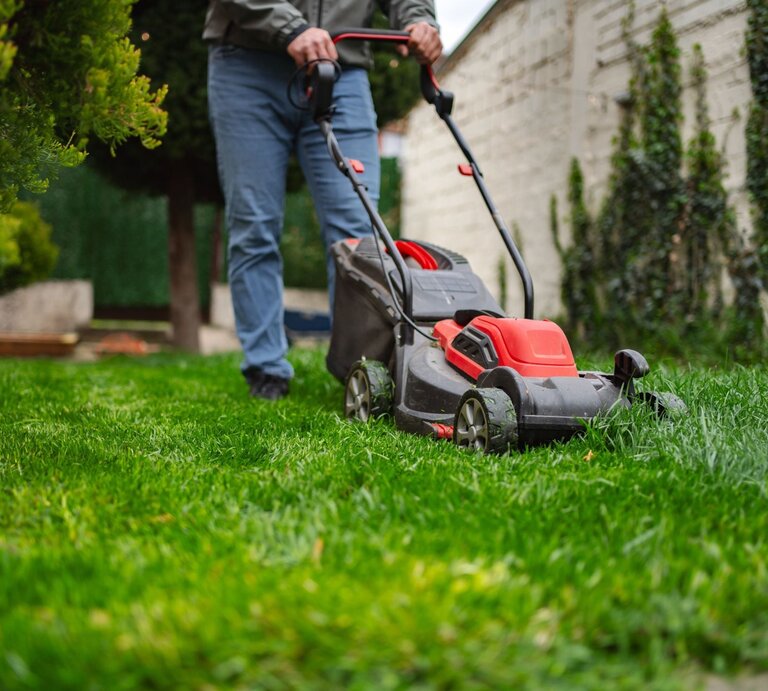 Eine Person in grauer Jacke und blauen Jeans mäht mit einem roten und schwarzen Rasenmäher grünes Gras in einem Garten mit Bäumen. | © Shutterstock/Magic Lens