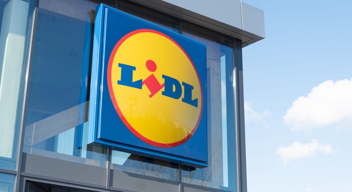  Das Bild zeigt das Logo des Einzelhandelsunternehmens Lidl an der Außenfassade eines Gebäudes. Das Logo ist in den charakteristischen Unternehmensfarben Blau und Gelb gehalten, mit einem roten Akzent, der den Teil des „i“ im Wort „Lidl“ darstellt. Der Hintergrund ist ein klarer Himmel mit einigen Wolken, was auf einen Standort des Geschäfts bei Tageslicht hinweist.