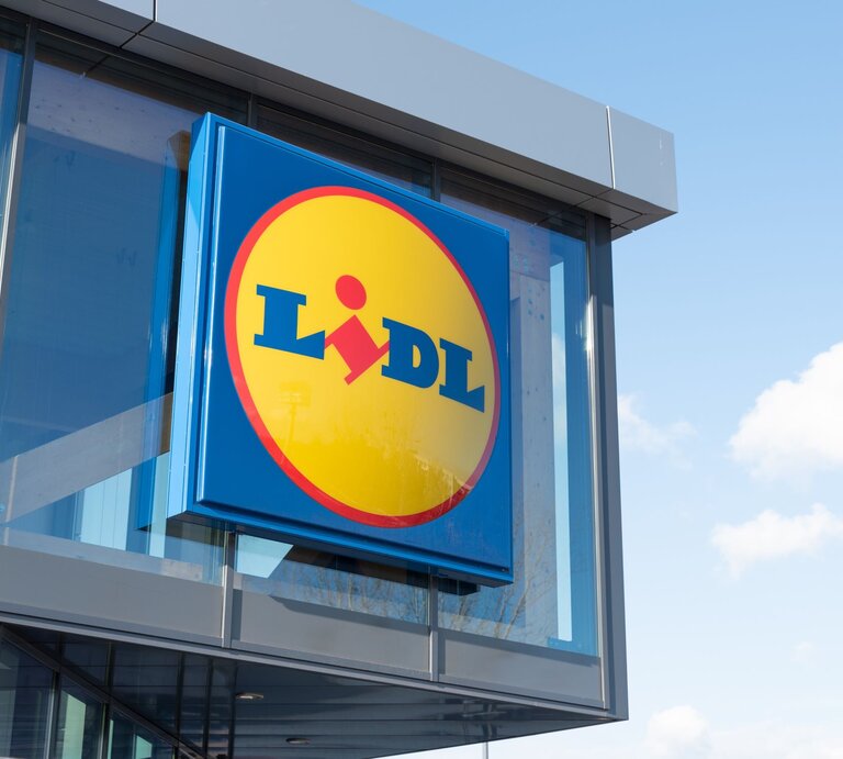  Das Bild zeigt das Logo des Einzelhandelsunternehmens Lidl an der Außenfassade eines Gebäudes. Das Logo ist in den charakteristischen Unternehmensfarben Blau und Gelb gehalten, mit einem roten Akzent, der den Teil des „i“ im Wort „Lidl“ darstellt. Der Hintergrund ist ein klarer Himmel mit einigen Wolken, was auf einen Standort des Geschäfts bei Tageslicht hinweist. | © AdobeStock/Proxima Studio