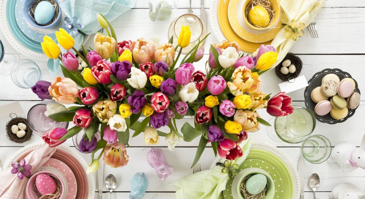 Bunte Tulpen in Gelb, Rot, Rosa und Lila schmücken eine festlich gedeckte Ostertafel mit pastellfarbenem Geschirr und verzierten Ostereiern.