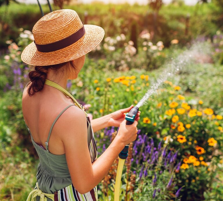 Junge Frau mit Strohhut gießt an einem sonnigen Tag bunte Sommerblumen im üppigen Garten mit einem Wasserschlauch. | © Shutterstock/Mariia Boiko