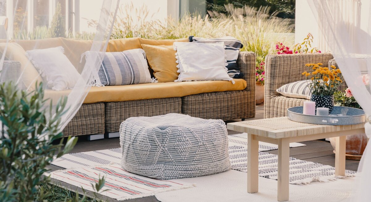 Gemütliche Terrasse mit Rattansofa, Holztisch und gemusterten Outdoorteppichen in Beige, Weiß und Rot, umgeben von Pflanzen