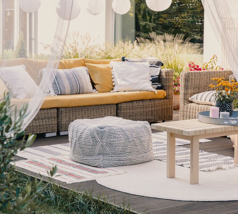 Gemütliche Terrasse mit Rattansofa, Holztisch und gemusterten Outdoorteppichen in Beige, Weiß und Rot, umgeben von Pflanzen | © Shutterstock/Ground Picture