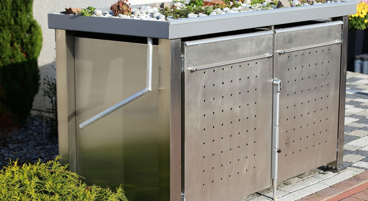 Moderne Mülltonnenbox aus Edelstahl mit bepflanztem Gründach, seitlichem Griff und perforierten Türen auf gepflastertem Untergrund.