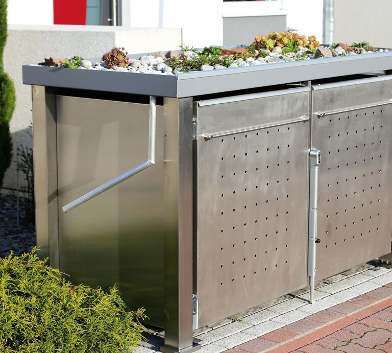 Moderne Mülltonnenbox aus Edelstahl mit bepflanztem Gründach, seitlichem Griff und perforierten Türen auf gepflastertem Untergrund. | © AdobeStock/ U. J. Alexander