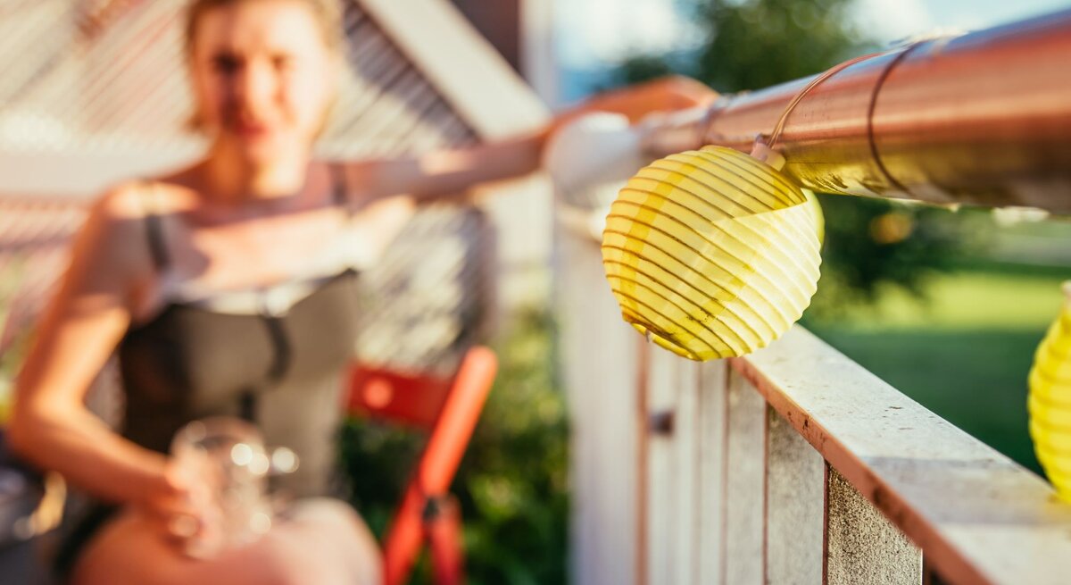 Junge Frau mit verschwommenem Gesicht in Sommerkleidung genießt die Sonne auf einem Balkon mit gelben Laternen.