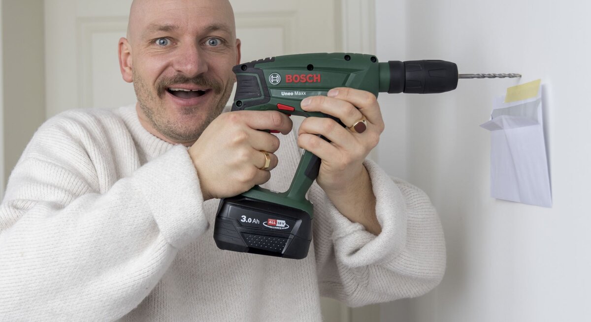 Ein fröhlicher Mann mit blauem Blick und Schnurrbart bohrt mit einer Bosch-Bohrmaschine nahe eines angeklebten Umschlags in eine Wand.