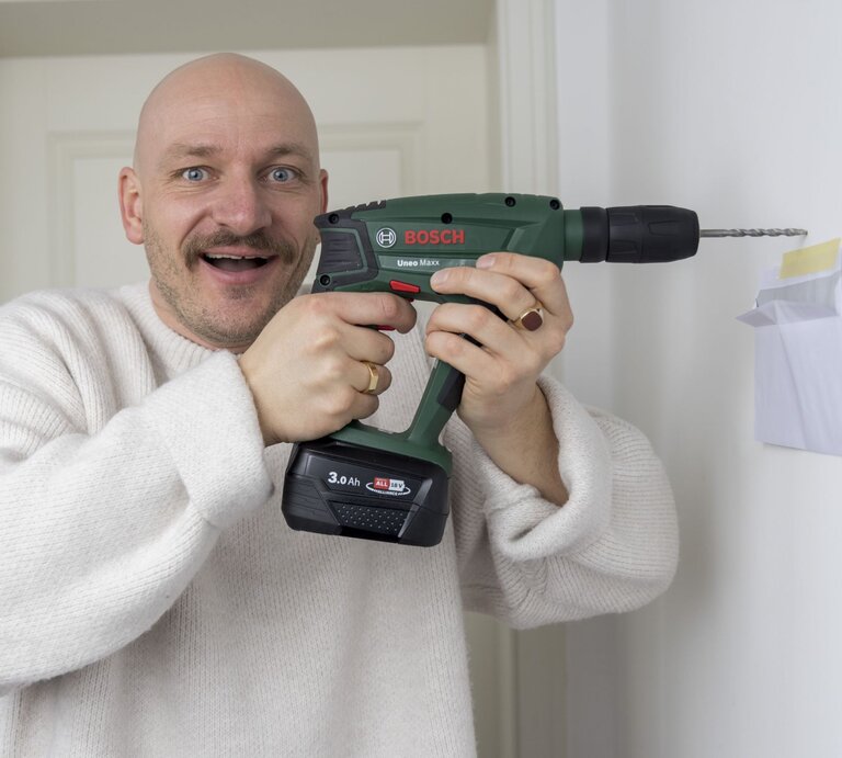 Ein fröhlicher Mann mit blauem Blick und Schnurrbart bohrt mit einer Bosch-Bohrmaschine nahe eines angeklebten Umschlags in eine Wand. | © BILD der FRAU 