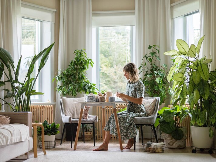 Eine Frau mit schulterlangem blondem Haar sitzt barfuß an einem Tisch in einem lichtdurchfluteten Raum mit großen, grünen Zimmerpflanzen. | © Shutterstock/Kostikova Natalia