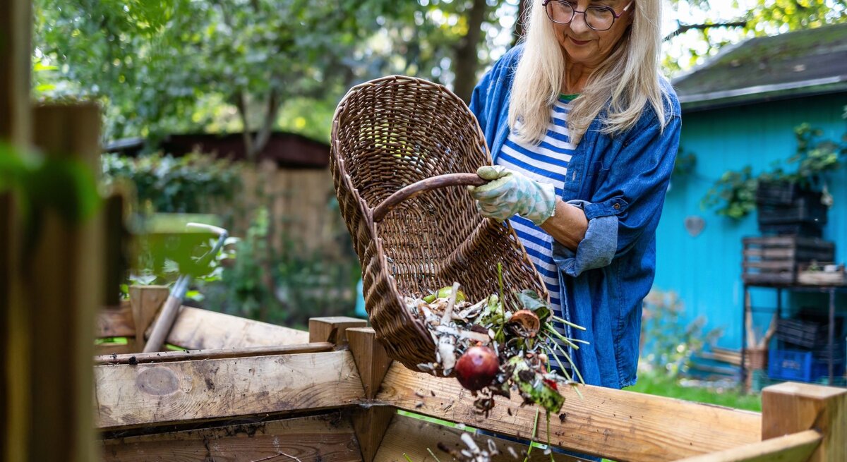 Eine ältere Frau mit blondem Haar und Brille leert lächelnd einen Korb mit Gartenabfällen in einen Kompostbehälter in grünem Garten.