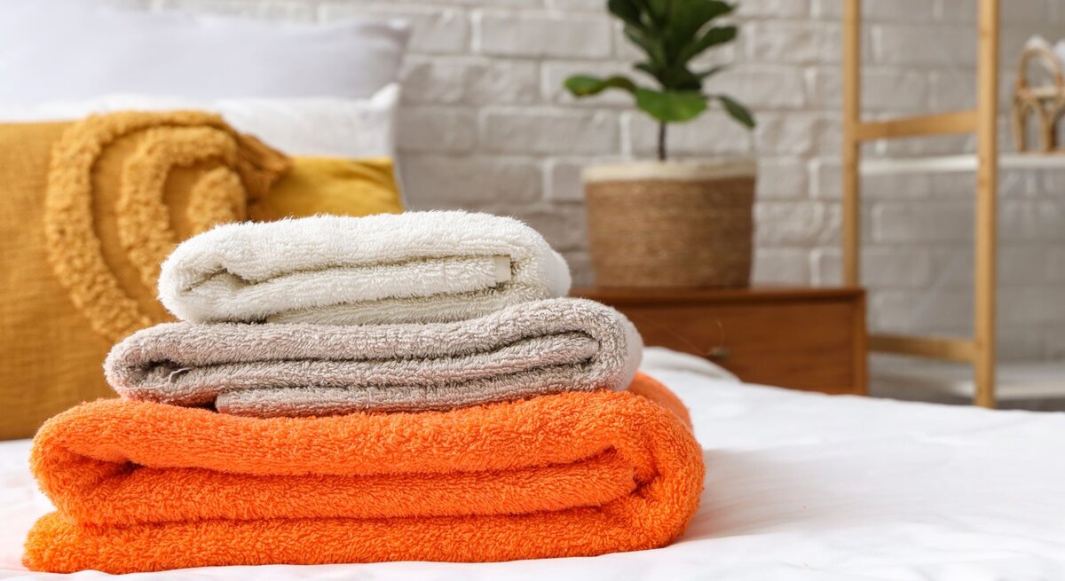 Drei gefaltete Handtücher in Weiß, Beige und Orange liegen gestapelt auf einem Bett, im Hintergrund eine Pflanze vor weißer Backsteinwand.