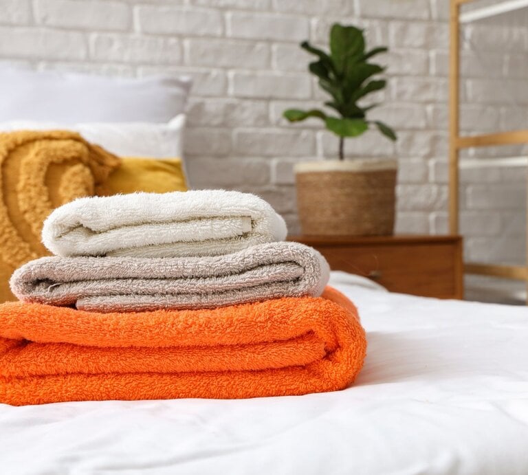 Drei gefaltete Handtücher in Weiß, Beige und Orange liegen gestapelt auf einem Bett, im Hintergrund eine Pflanze vor weißer Backsteinwand. | © Shutterstock/Pixel-Shot