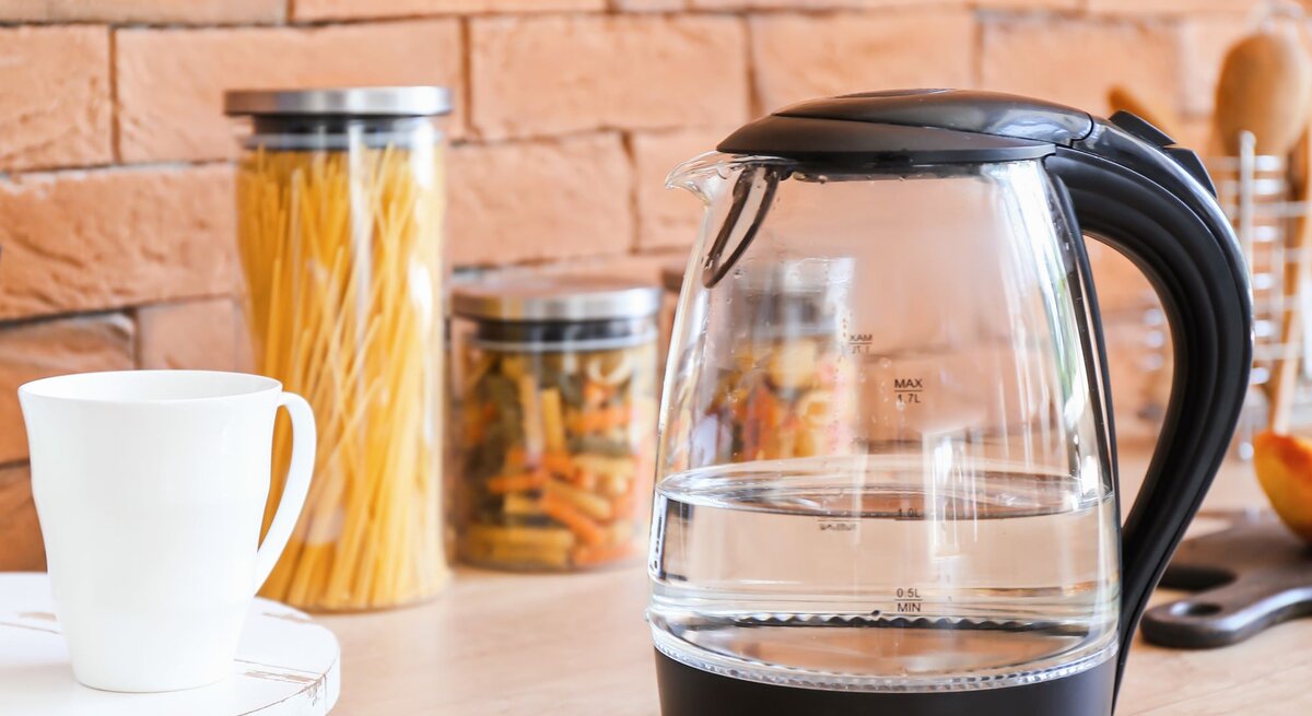 Ein transparenter Wasserkocher mit schwarzem Griff steht auf einer Holzarbeitsplatte, daneben eine weiße Tasse und Gläser mit Nudeln vor einer Ziegelwand.
