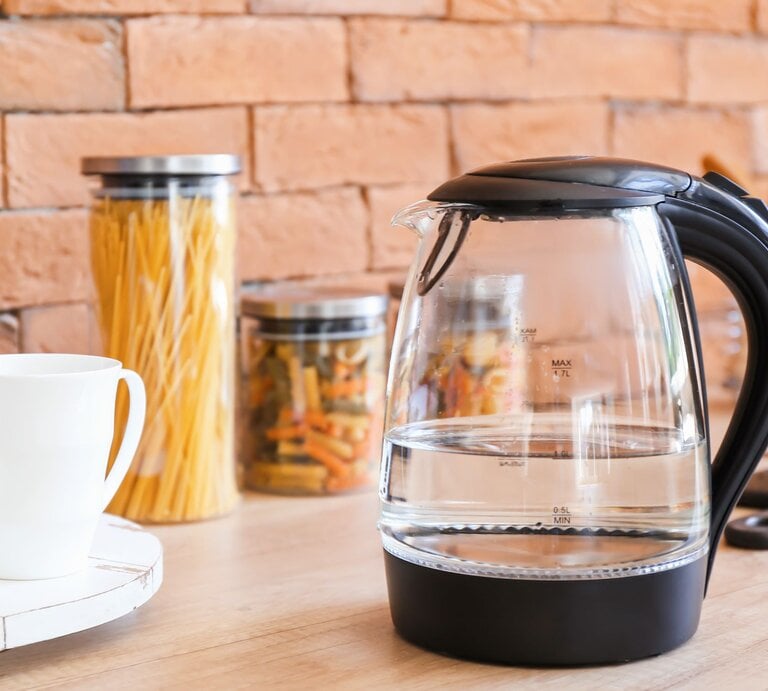 Ein transparenter Wasserkocher mit schwarzem Griff steht auf einer Holzarbeitsplatte, daneben eine weiße Tasse und Gläser mit Nudeln vor einer Ziegelwand. | © Adobe Stock/Pixel-Shot