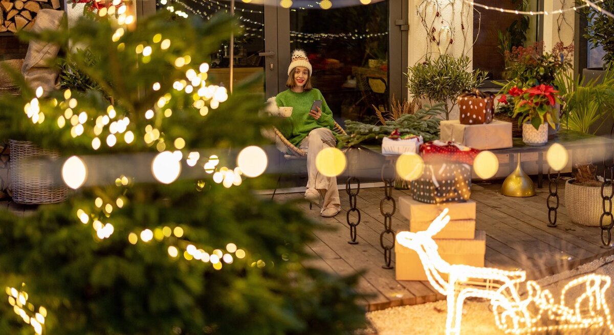 Eine junge Frau mit weißer Mütze und grünem Pullover sitzt lächelnd auf einer Terrasse, umgeben von Weihnachtsdeko und Geschenken.