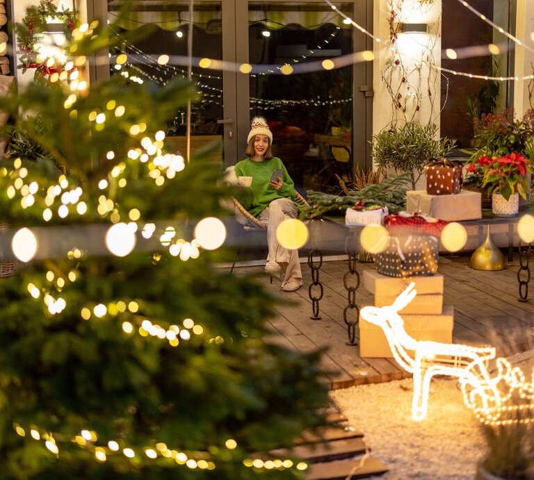 Eine junge Frau mit weißer Mütze und grünem Pullover sitzt lächelnd auf einer Terrasse, umgeben von Weihnachtsdeko und Geschenken. | © Shutterstock/RossHelen