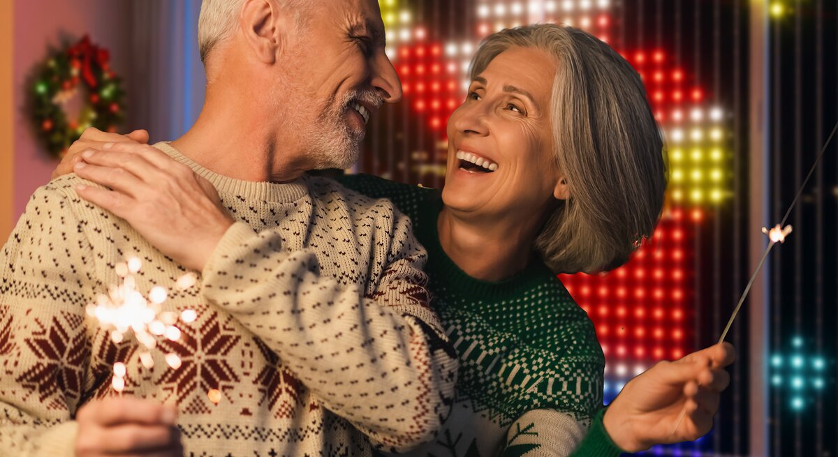 Ein älteres Paar in Weihnachtspullovern lacht sich liebevoll an und hält Wunderkerzen, im Hintergrund festliche Lichterdeko.