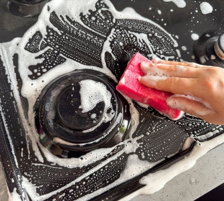 Eine Hand mit heller Haut reinigt mit einem pinken Schwamm und Seifenschaum eine schwarze Herdplatte mit glänzenden Kochfeldern. | © Shutterstock/RomanR