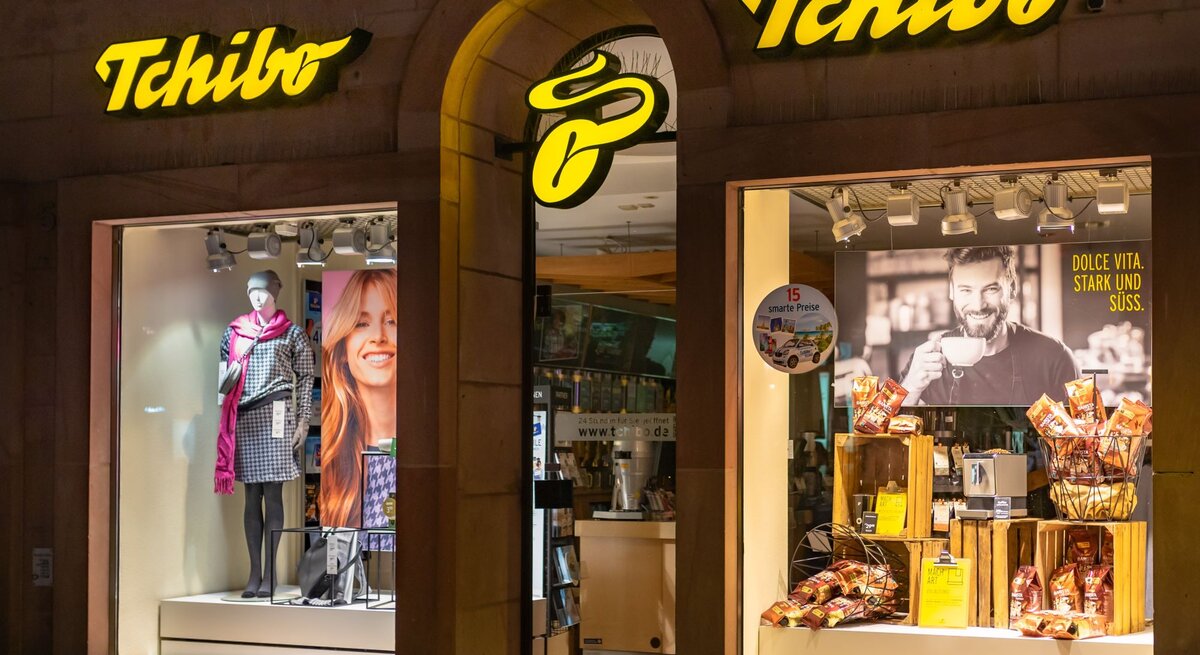 Tchibo-Schaufenster bei Nacht mit Modepuppe in karierter Jacke, rotem Schal und Werbung für Kaffee mit lächelndem Mann im Hintergrund.