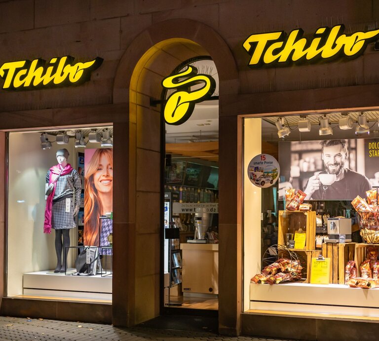 Tchibo-Schaufenster bei Nacht mit Modepuppe in karierter Jacke, rotem Schal und Werbung für Kaffee mit lächelndem Mann im Hintergrund. | © Shutterstock/vulcano