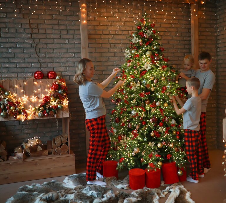 Eine lächelnde Familie schmückt in karierten Schlafanzügen einen leuchtenden Weihnachtsbaum vor einer Backsteinwand mit Lichterketten. | © GettyImages/Natalia Lebedinskaia