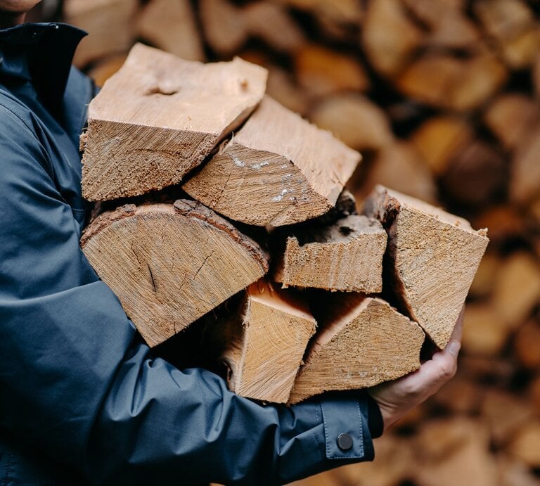 Eine Person in blauer Jacke trägt mehrere grob gespaltene Holzscheite vor einem Stapel Holz im Hintergrund. | © AdobeStock/VK Studio