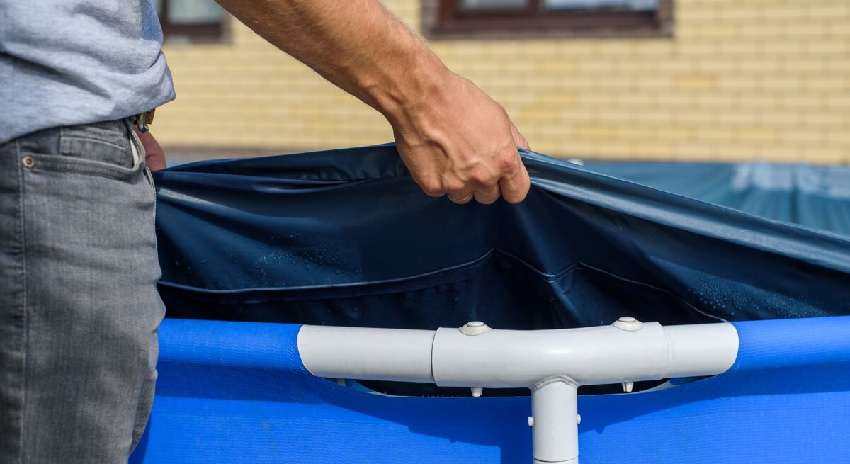 Ein Mann in grauem T-Shirt und Jeans spannt eine blaue Poolfolie über einen Aufstellpool vor einem gelben Backsteingebäude.
