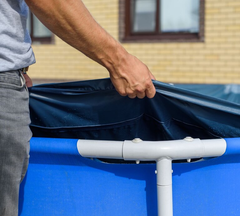 Ein Mann in grauem T-Shirt und Jeans spannt eine blaue Poolfolie über einen Aufstellpool vor einem gelben Backsteingebäude. | © AdobeStock/penyushkin
