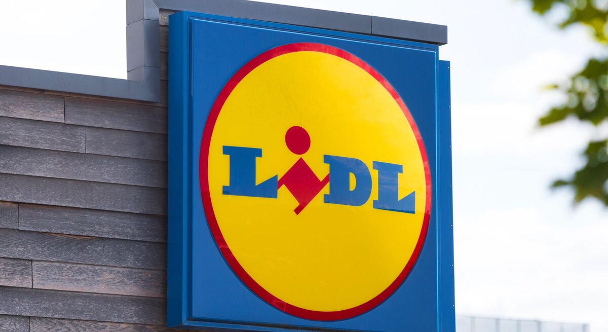 Das Bild zeigt das Firmenschild eines Lidl-Marktes. Im Mittelpunkt steht das markante Firmenlogo auf einem blauen Hintergrund, bestehend aus einem gelben Kreis mit einem roten Rand, in dem der Name "Lidl" in blauen Buchstaben steht. Innerhalb des gelben Kreises befindet sich das stilisierte Bild einer Person, die einen Einkaufswagen schiebt. Das Schild ist an der Seite eines Gebäudes mit einer hölzernen Fassade befestigt. Im Hintergrund sieht man einen leicht bewölkten Himmel und das Laub eines Baumes in der Ecke oben rechts. Das Bild wirkt klar und modern, typisch für die Außengestaltung eines Supermarktes der Kette.