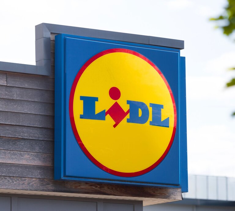 Das Bild zeigt das Firmenschild eines Lidl-Marktes. Im Mittelpunkt steht das markante Firmenlogo auf einem blauen Hintergrund, bestehend aus einem gelben Kreis mit einem roten Rand, in dem der Name "Lidl" in blauen Buchstaben steht. Innerhalb des gelben Kreises befindet sich das stilisierte Bild einer Person, die einen Einkaufswagen schiebt. Das Schild ist an der Seite eines Gebäudes mit einer hölzernen Fassade befestigt. Im Hintergrund sieht man einen leicht bewölkten Himmel und das Laub eines Baumes in der Ecke oben rechts. Das Bild wirkt klar und modern, typisch für die Außengestaltung eines Supermarktes der Kette. | © Adobe Stock/ Tobias Arhelger