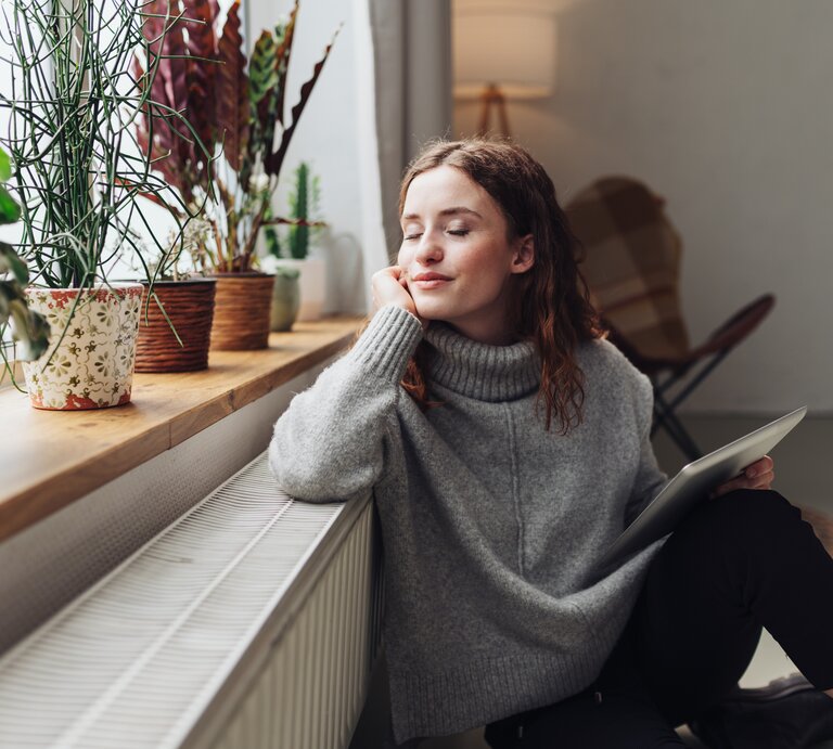 Junge Frau mit langen braunen Haaren im grauen Pullover sitzt verträumt an der Heizung am Fenster, umgeben von Zimmerpflanzen. | © AdobeStock/contrastwerkstatt