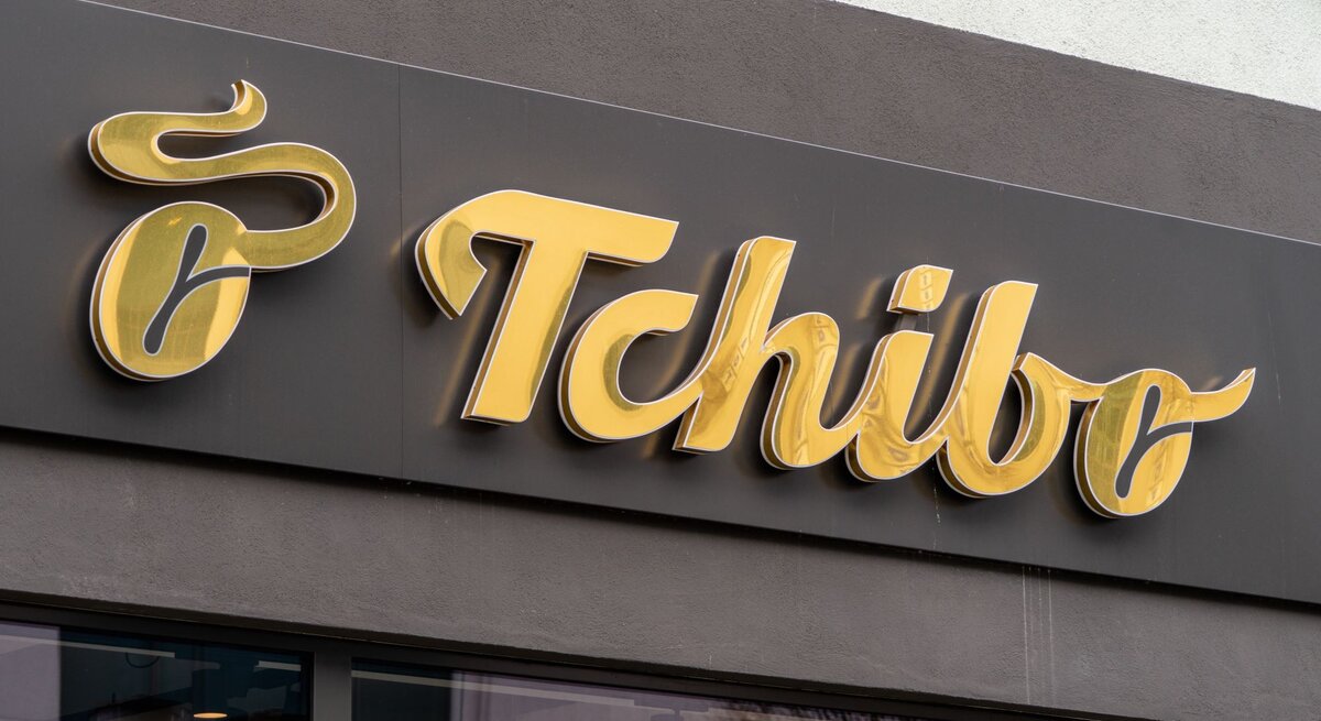 Goldenes Tchibo-Logo auf schwarzem Hintergrund an einer Hausfassade, unterhalb Fenster mit Spiegelung von Bäumen.