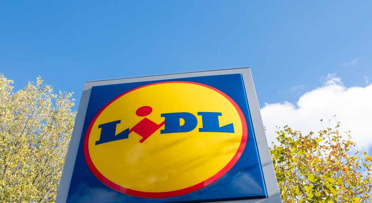  Das Bild zeigt das Logo von Lidl auf einem großen Schild, das gegen den Himmel und einige Baumkronen im Hintergrund steht. Das Logo besteht aus einem blauen Rechteck mit abgerundeten Ecken, in dem der Name "Lidl" in weißer Schrift steht.