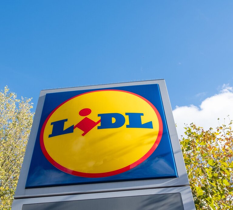  Das Bild zeigt das Logo von Lidl auf einem großen Schild, das gegen den Himmel und einige Baumkronen im Hintergrund steht. Das Logo besteht aus einem blauen Rechteck mit abgerundeten Ecken, in dem der Name "Lidl" in weißer Schrift steht. | © Adobe Stock/William