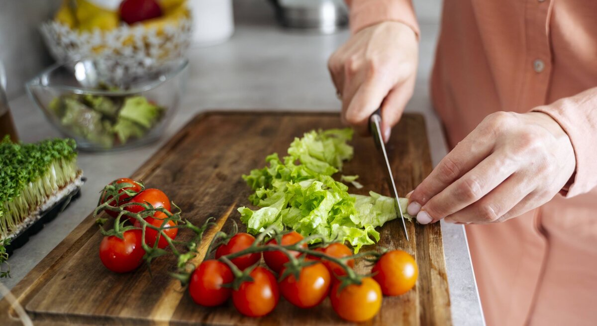 Eine Person in rosa Bluse schneidet auf einem Holzbrett frischen grünen Salat neben leuchtend roten Tomaten.