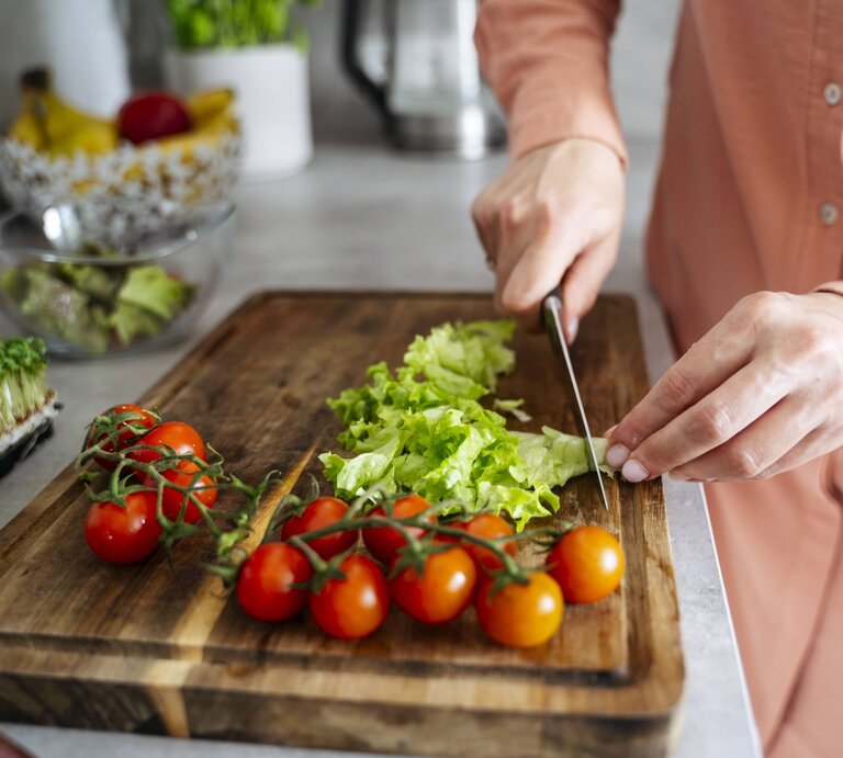 Eine Person in rosa Bluse schneidet auf einem Holzbrett frischen grünen Salat neben leuchtend roten Tomaten. | © IMAGO/Westend61