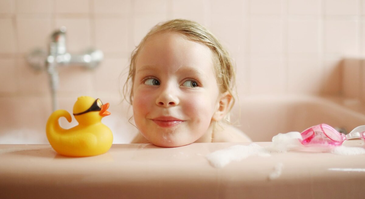 Ein blondes Kleinkind mit verschmitztem Lächeln schaut in einer rosafarbenen Badewanne neben einer gelben Gummiente hervor.