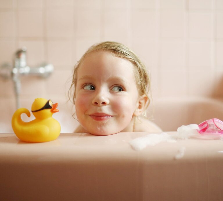 Ein blondes Kleinkind mit verschmitztem Lächeln schaut in einer rosafarbenen Badewanne neben einer gelben Gummiente hervor. | © Getty Images/Catherine Delahaye