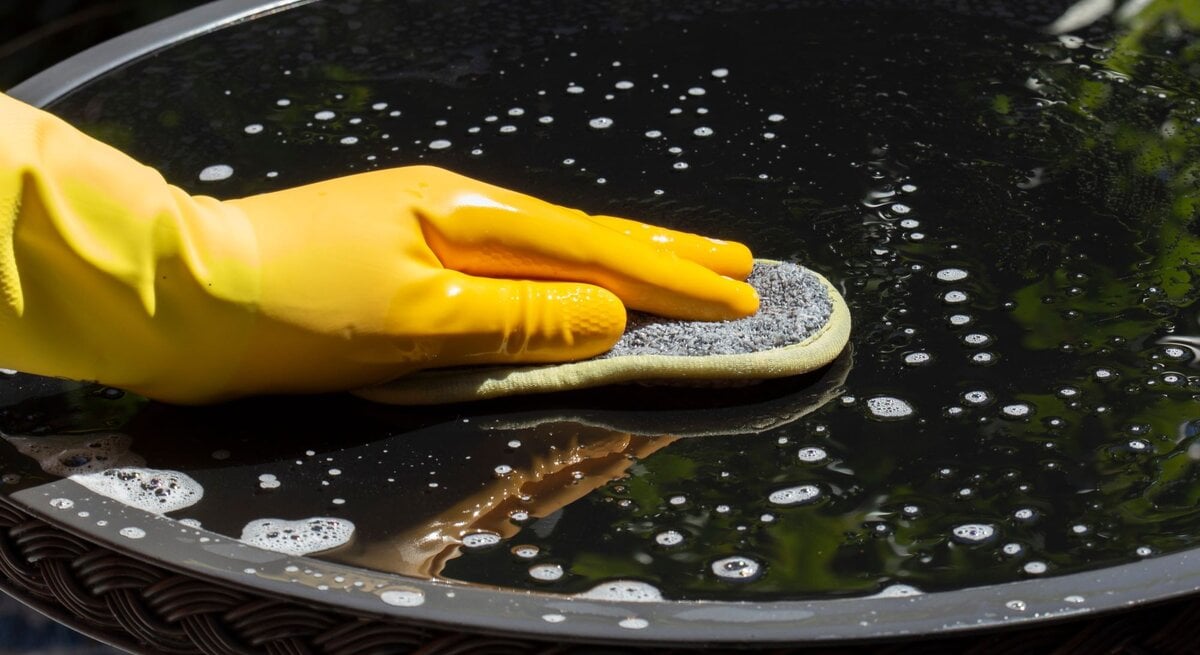 Eine Hand mit gelbem Gummihandschuh reinigt mit einem Schwamm einen nassen Glastisch, auf dem Seifenblasen schimmern.