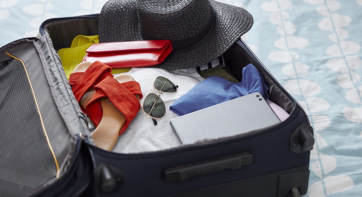 Ein offener Koffer auf einem gemusterten Bett, gefüllt mit einem schwarzen Hut, roten Sandalen, Sonnenbrille, Tablet und Kleidung.