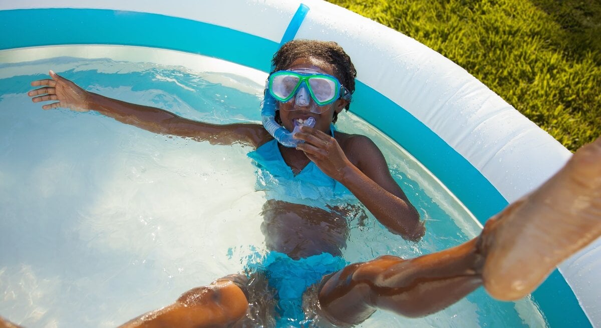 Ein fröhliches Mädchen in blauer Badebekleidung und Taucherbrille planscht mit ausgestreckten Armen im aufblasbaren Pool im Garten.